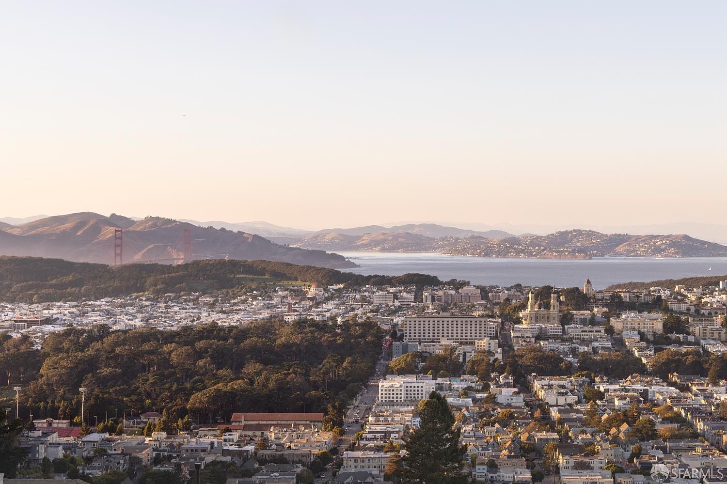 This aerial view showcases a sprawling cityscape with a mix of residential and commercial buildings, interspersed with lush greenery. In the distance, a bridge and the bay provide a scenic backdrop, creating a sense of urban living combined with natural beauty. The overall impression is one of a vibrant and desirable location.