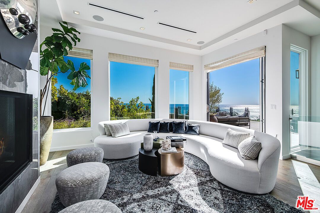 This sophisticated living room features a large, curved white sofa set against a backdrop of floor-to-ceiling windows that offer a stunning, unobstructed view of the ocean. The space is anchored by a textured dark gray area rug and a modern two-piece coffee table, complemented by plush gray ottomans and a tall indoor plant. The bright, airy atmosphere is enhanced by recessed lighting and a sleek fireplace, creating a luxurious and serene coastal living environment.