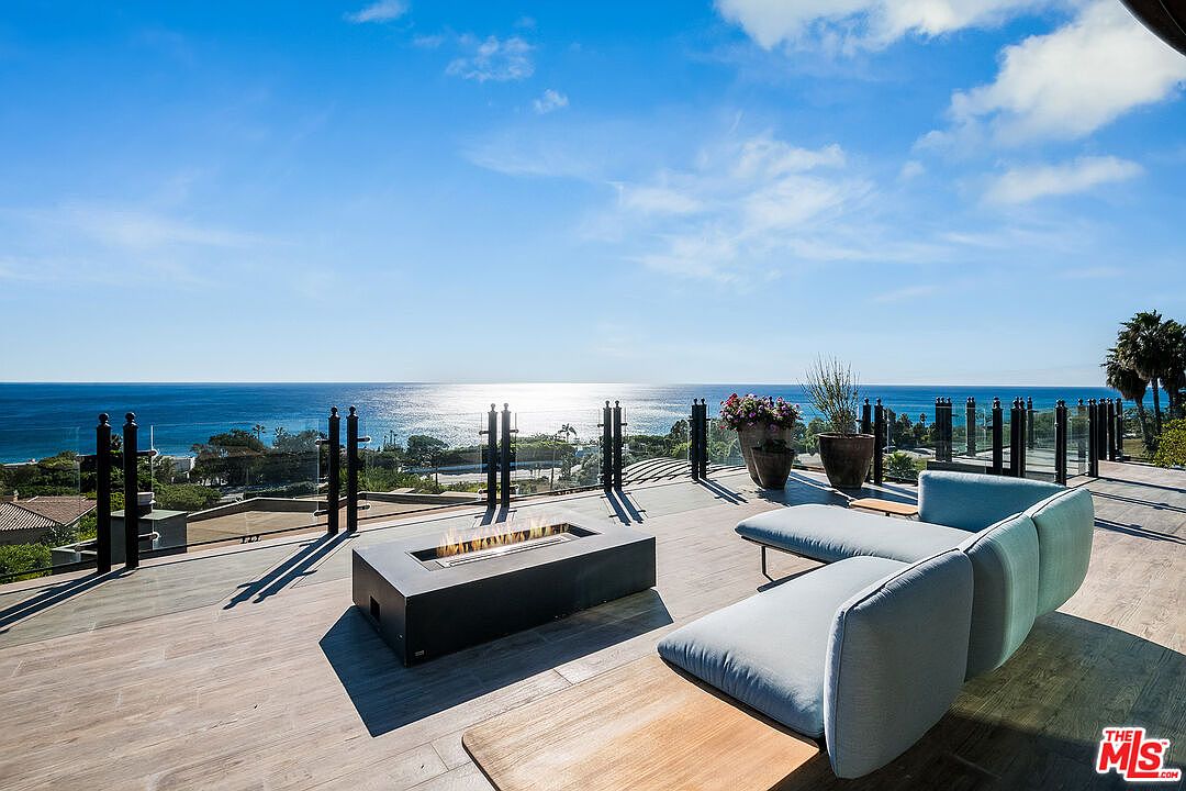 This expansive, modern outdoor deck offers a breathtaking, unobstructed panoramic view of the ocean under a bright, sunny sky. The space is elegantly furnished with a contemporary light blue sectional sofa and a sleek, rectangular fire pit, creating an ideal setting for luxury relaxation and entertaining. The clean lines of the glass railing and the wood-look tile flooring emphasize the sophisticated, high-end coastal aesthetic of this property.