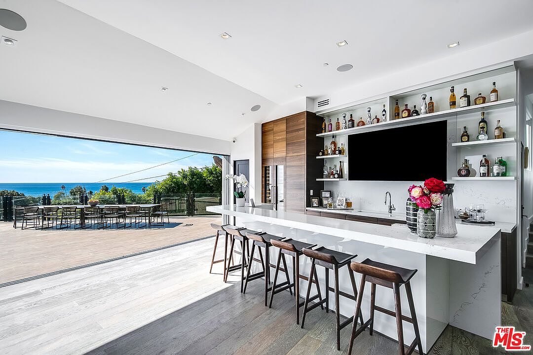 This sophisticated, open-concept kitchen and bar area features a sleek white quartz waterfall island with dark wood bar stools, seamlessly transitioning to an expansive outdoor deck through floor-to-ceiling sliding glass doors. The space is highlighted by a custom built-in bar with illuminated shelving, a large television, and a stunning panoramic view of the ocean. The modern design aesthetic combines warm wood cabinetry with clean, minimalist lines to create a luxurious and airy atmosphere.