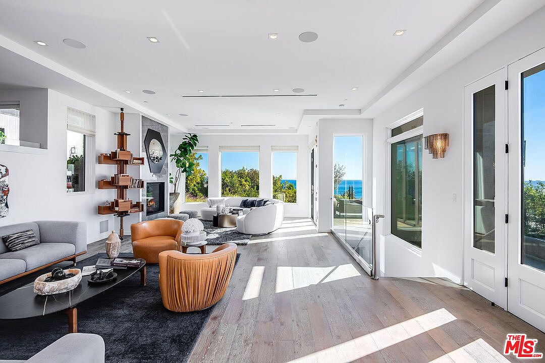 This bright and airy living room features a sophisticated open-concept design with light hardwood floors and expansive windows that frame a stunning ocean view. The space is furnished with a mix of modern grey and warm tan leather seating, centered around a sleek black coffee table and a unique, floor-to-ceiling wooden shelving unit. The room exudes a luxurious, contemporary coastal aesthetic, enhanced by recessed lighting and a minimalist fireplace.