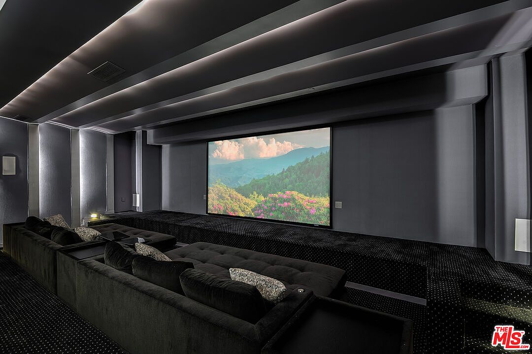 This luxurious home theater features a tiered seating arrangement with plush, dark velvet sofas and integrated side tables, creating an immersive viewing experience. The room is designed with a sophisticated, dark aesthetic, highlighted by recessed cove lighting and a large, high-definition projection screen displaying a scenic landscape. The elevated platform and tiered layout provide optimal sightlines, emphasizing a premium, cinematic atmosphere perfect for private entertainment.