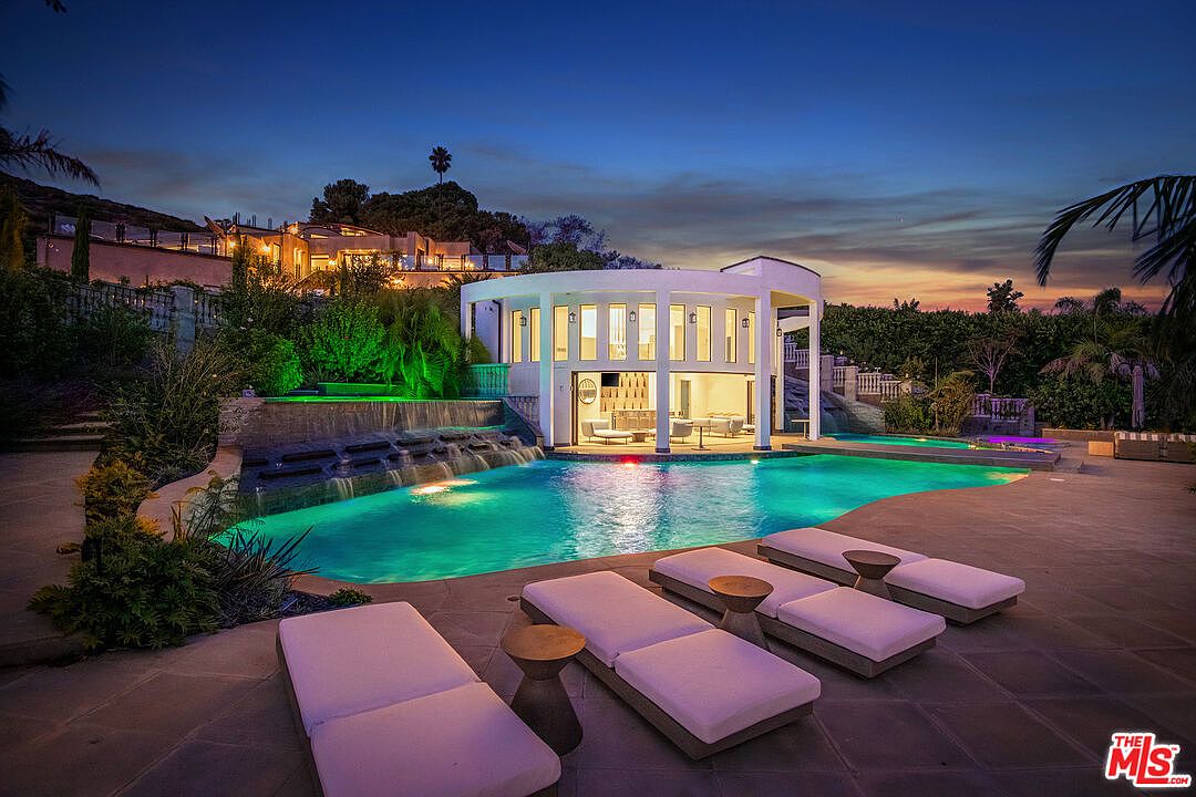 This stunning twilight shot captures a luxurious backyard oasis featuring a large, illuminated swimming pool with a cascading waterfall feature. A modern, circular pool house with floor-to-ceiling glass walls serves as a focal point, complemented by sleek lounge chairs arranged on the expansive stone patio. The scene exudes a high-end, resort-like atmosphere, perfectly framed to showcase the property's sophisticated outdoor entertainment space.