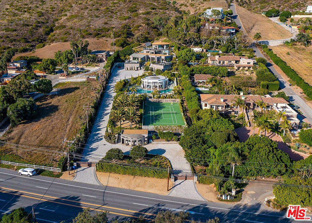 This high-angle aerial view captures a sprawling, multi-level estate nestled into a hillside, featuring a prominent circular pool house, a private tennis court, and extensive landscaped grounds. The property is accessed by a long, paved driveway leading from the main road, showcasing a blend of modern and traditional architectural styles across several structures. The surrounding terrain is characterized by dry, rolling hills, emphasizing the secluded and expansive nature of this luxury residential compound.