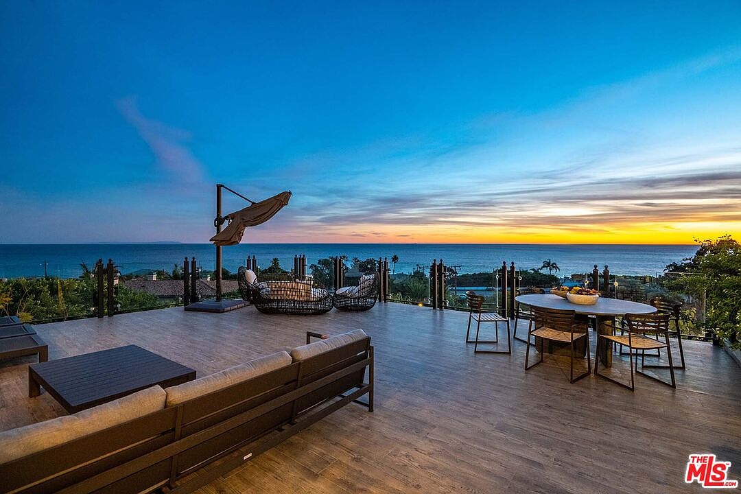 This expansive, elevated deck offers a breathtaking panoramic view of the ocean at sunset, creating a serene and luxurious atmosphere. The space is furnished with a modern outdoor sofa, a dining set, and lounge chairs, making it an ideal setting for alfresco entertaining. The wood-look flooring and sleek glass railing emphasize the contemporary design while ensuring an unobstructed connection to the horizon.