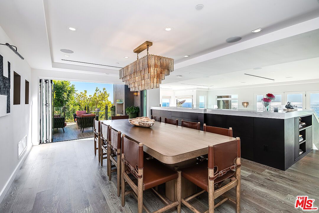 This elegant dining room features a large wooden table surrounded by leather-upholstered chairs, anchored by a striking, multi-tiered crystal chandelier. The space seamlessly transitions to an outdoor deck through expansive folding glass doors, creating an airy, indoor-outdoor living experience. The room is finished with light-toned hardwood flooring and a clean, modern aesthetic that overlooks a bright, open-concept living area.