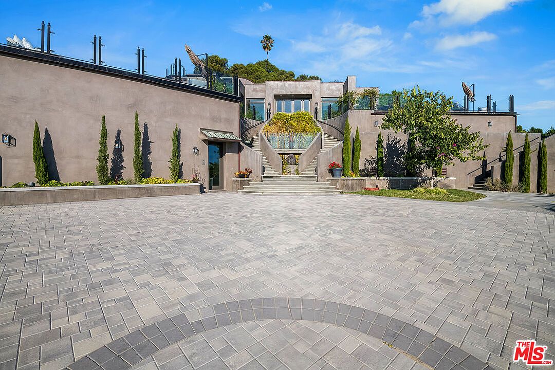 This grand, modern estate features a sprawling paved motor court leading to a dramatic, symmetrical staircase that ascends to the main entrance. The exterior is finished in a smooth, neutral stucco, accented by manicured cypress trees and tiered landscaping that creates a sophisticated, architectural aesthetic. The perspective is a wide, eye-level shot that emphasizes the scale and imposing presence of the property's facade.