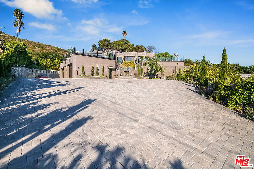 This expansive front exterior view showcases a grand, modern estate approached by a massive, meticulously paved stone driveway. The multi-level structure features clean, contemporary lines, a prominent central staircase leading to the main entrance, and integrated garage bays on the left. Surrounded by lush landscaping and tall, slender cypress trees, the property exudes a sense of luxury, privacy, and architectural sophistication under a clear blue sky.