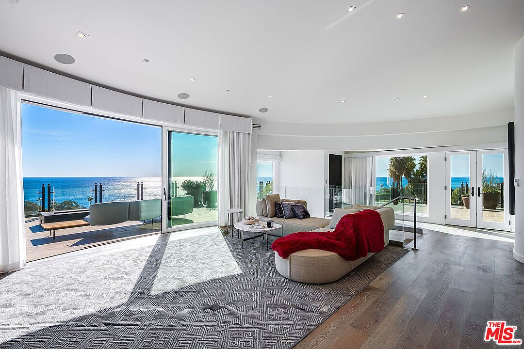 This expansive, light-filled living room features a modern, open-concept design with floor-to-ceiling glass sliding doors that seamlessly blend the indoor space with a stunning oceanfront deck. The room is anchored by a large, curved sectional sofa draped with a vibrant red throw, set against a backdrop of minimalist white walls and rich hardwood flooring. The panoramic coastal views and high-end finishes create an atmosphere of sophisticated luxury and serene relaxation.
