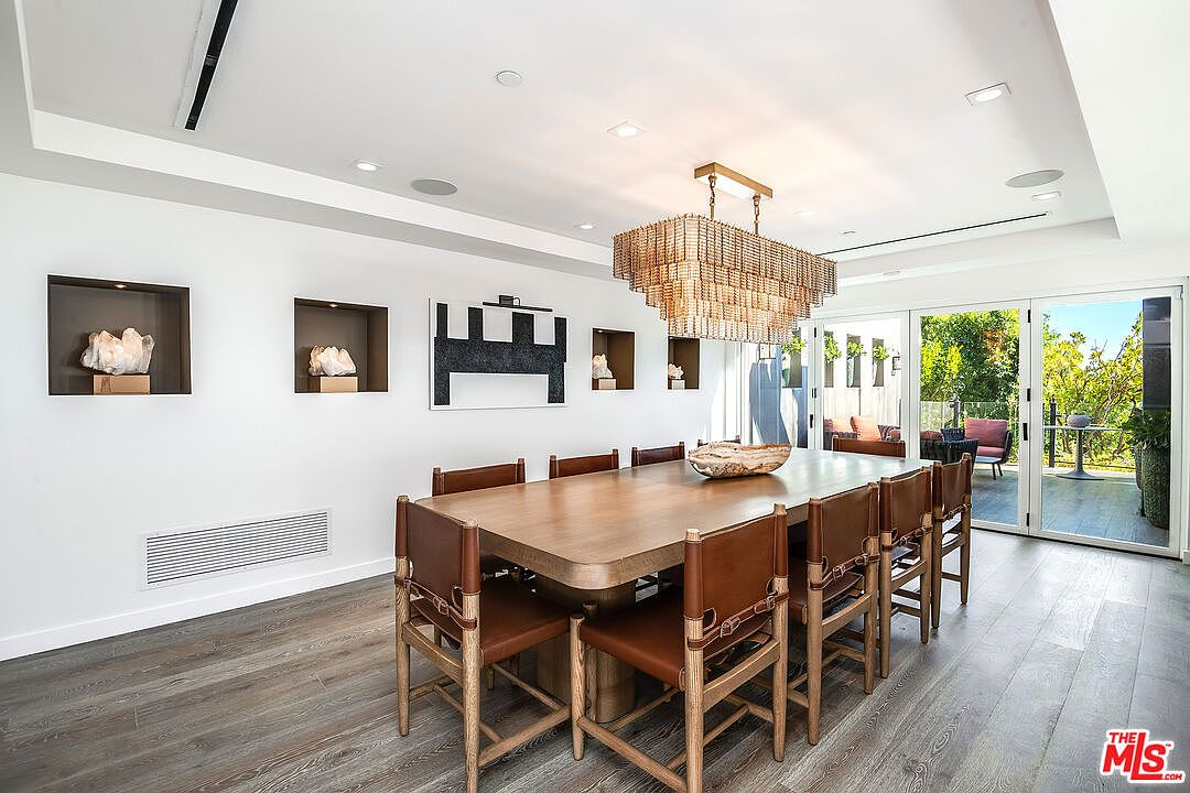 This elegant dining room features a large, warm-toned wooden table surrounded by eight leather-upholstered chairs with a distinct rustic-modern aesthetic. A grand, tiered beaded chandelier serves as the focal point above the table, while recessed wall niches display decorative crystal sculptures. The space is bright and airy, with floor-to-ceiling glass doors that seamlessly connect the interior to an outdoor patio area.