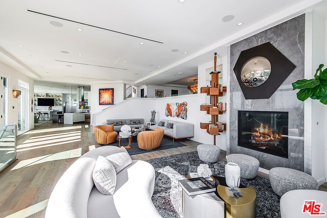 This expansive, open-concept living area showcases a sophisticated modern design with a striking floor-to-ceiling fireplace featuring a unique hexagonal mirror accent. The space is divided into two distinct seating areas, highlighted by a plush, curved white sofa in the foreground and a warm, earthy-toned arrangement in the background, all set against light hardwood floors. The room is bathed in natural light, creating an airy and luxurious atmosphere that flows seamlessly into the adjacent kitchen.