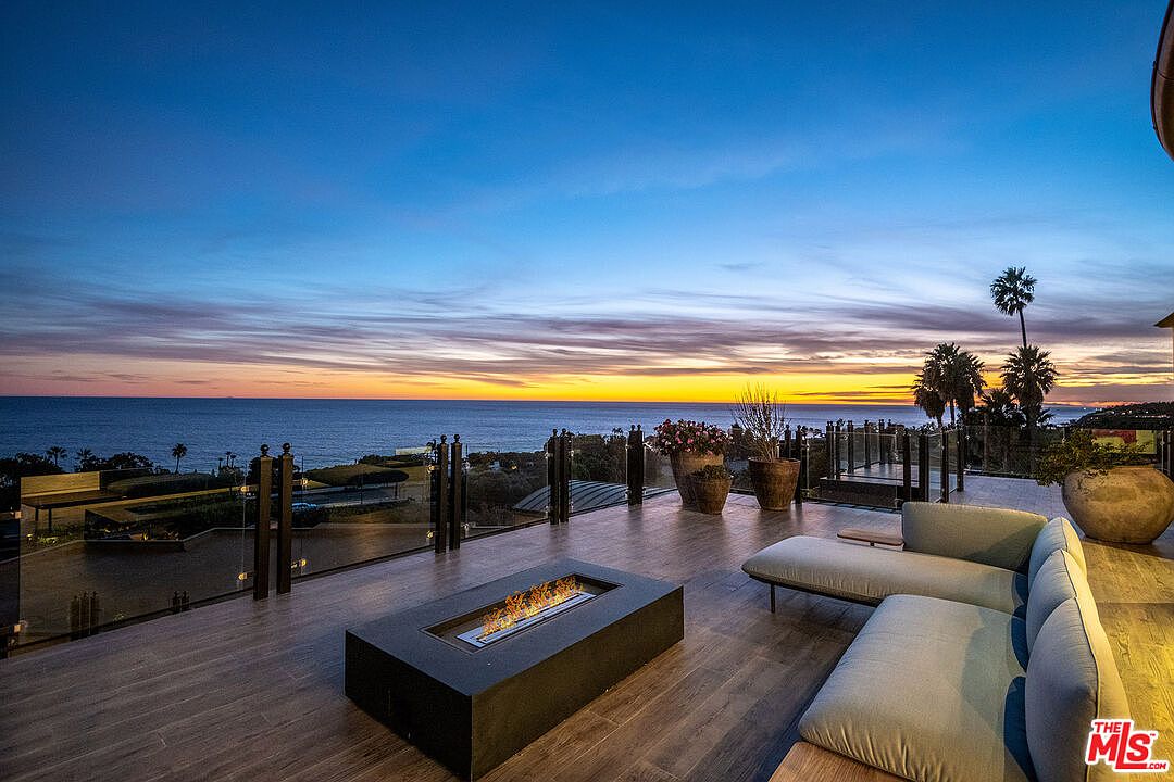 This stunning outdoor deck offers a panoramic, elevated view of the ocean at sunset, featuring a sleek, modern fire pit as a central focal point. The space is furnished with comfortable, contemporary lounge seating, perfect for relaxing while taking in the vibrant colors of the twilight sky. The glass railing ensures an unobstructed view, creating a seamless connection between the luxurious living space and the coastal horizon.