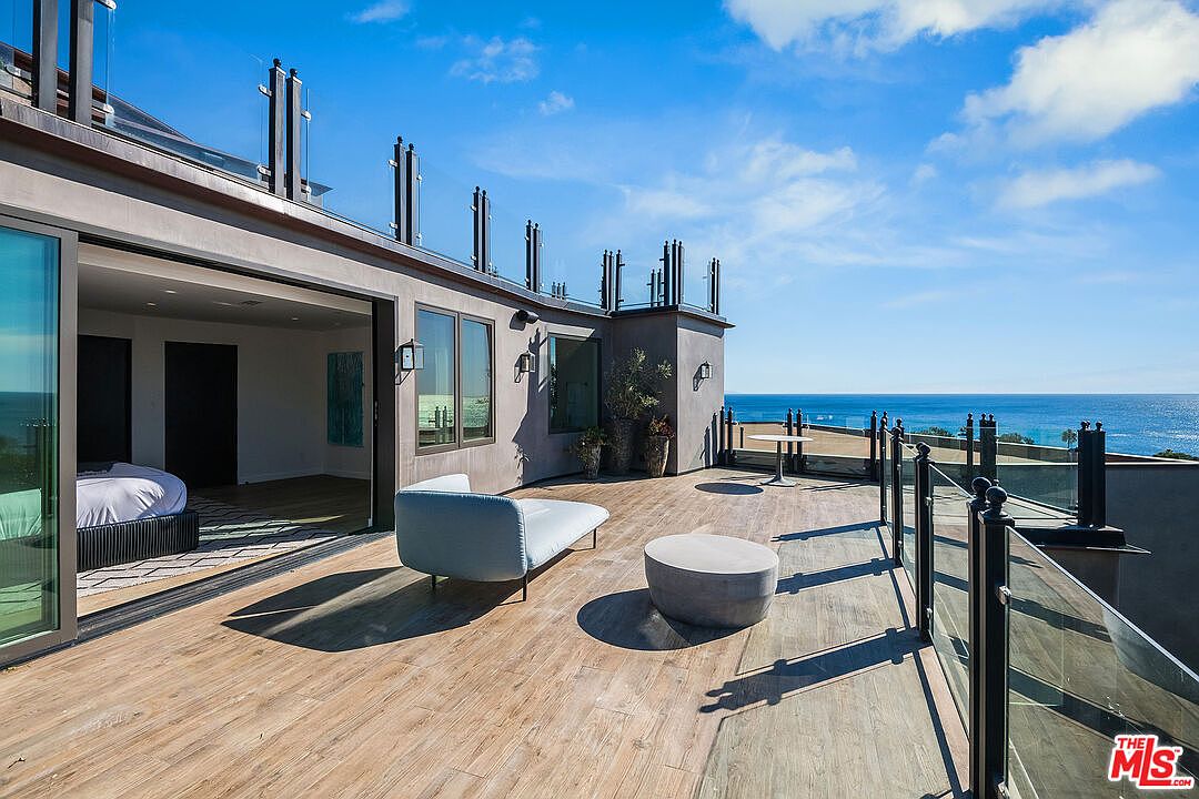 This expansive, modern balcony offers a seamless indoor-outdoor living experience with large sliding glass doors opening from a bedroom onto a spacious wood-look deck. The area is furnished with a sleek chaise lounge and a minimalist concrete coffee table, all framed by a contemporary glass railing that provides unobstructed, panoramic views of the ocean. The bright, sunny atmosphere and clean architectural lines create a luxurious and serene coastal retreat.