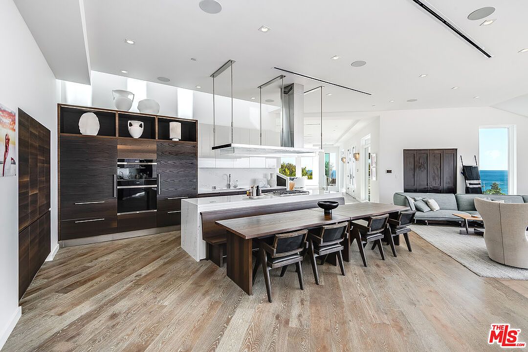 This expansive, modern open-concept kitchen features dark wood cabinetry, a large white marble-topped island, and an integrated dining table. The space is illuminated by recessed lighting and a sleek, suspended range hood, while light-toned hardwood floors provide a warm contrast to the dark cabinetry. A living area is visible in the background, creating a seamless flow that emphasizes the home's airy, contemporary design and ocean views.
