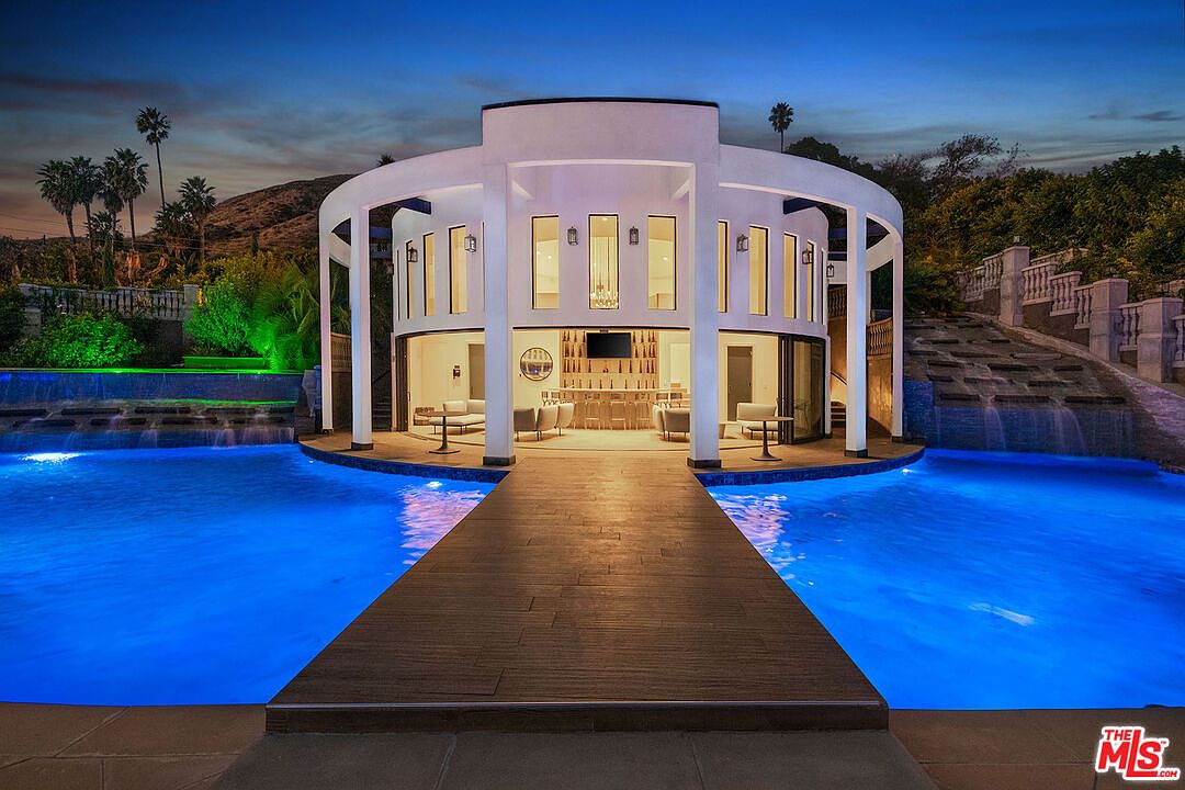 This stunning evening shot captures a luxurious, circular pool house pavilion situated in the center of a large, illuminated swimming pool. A wooden walkway leads directly to the pavilion, which features an open-air design, modern bar seating, and elegant architectural columns. The scene is enhanced by dramatic blue pool lighting and a cascading water feature in the background, creating a sophisticated and resort-like atmosphere.