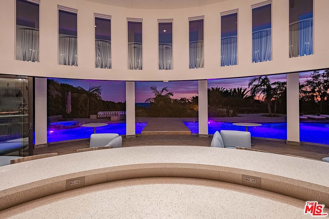 This sophisticated interior space features a curved, light-colored stone bar that overlooks a stunning, illuminated swimming pool at dusk. The room is defined by a series of floor-to-ceiling windows that create a seamless transition between the indoor living area and the vibrant outdoor landscape. The overall aesthetic is modern and luxurious, emphasizing a panoramic view of the sunset over the water.