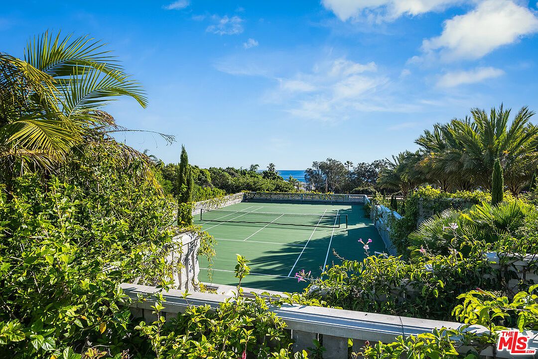 This elevated perspective showcases a private, well-maintained tennis court nestled within lush, tropical landscaping. The court features a classic green surface and is framed by vibrant greenery and palm trees, with a glimpse of the ocean visible in the distance. The scene conveys a sense of luxury, privacy, and an active outdoor lifestyle.