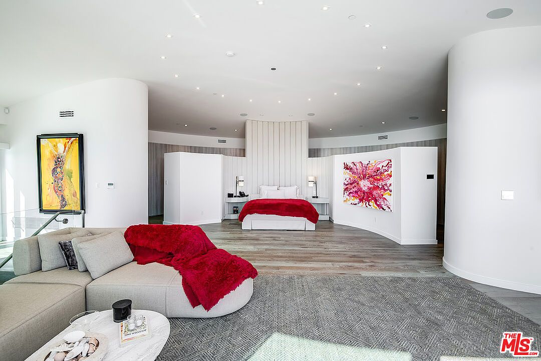 This expansive, circular primary bedroom features a contemporary open-concept design with a central bed dressed in a vibrant red throw. The room is anchored by a large, light-gray sectional sofa in the foreground and accented by bold, colorful abstract artwork on the curved white walls. The space is illuminated by recessed ceiling lights and offers a sophisticated, modern aesthetic with high-end finishes and a spacious, airy atmosphere.