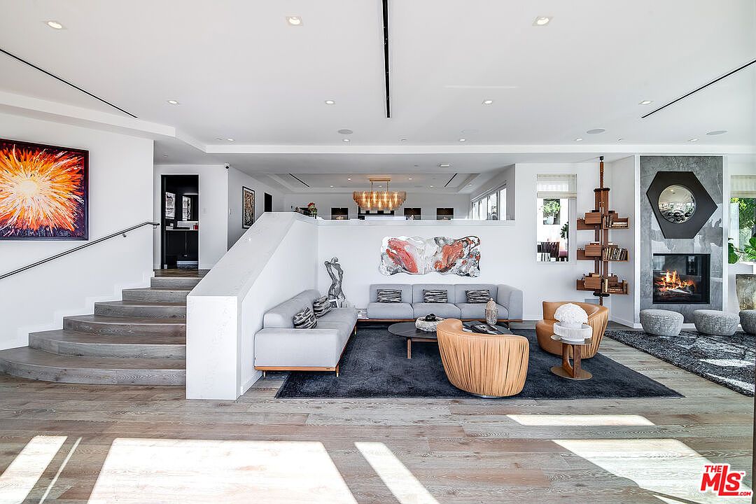 This expansive, open-concept living room showcases a sophisticated modern aesthetic with a sunken seating area defined by a plush dark rug and contemporary furniture. The space features a striking fireplace with a geometric surround, a floating wooden bookshelf, and a grand staircase leading to an upper level, all illuminated by recessed lighting and a distant chandelier. The neutral color palette, accented by vibrant abstract art, creates a luxurious and inviting atmosphere perfect for high-end entertaining.
