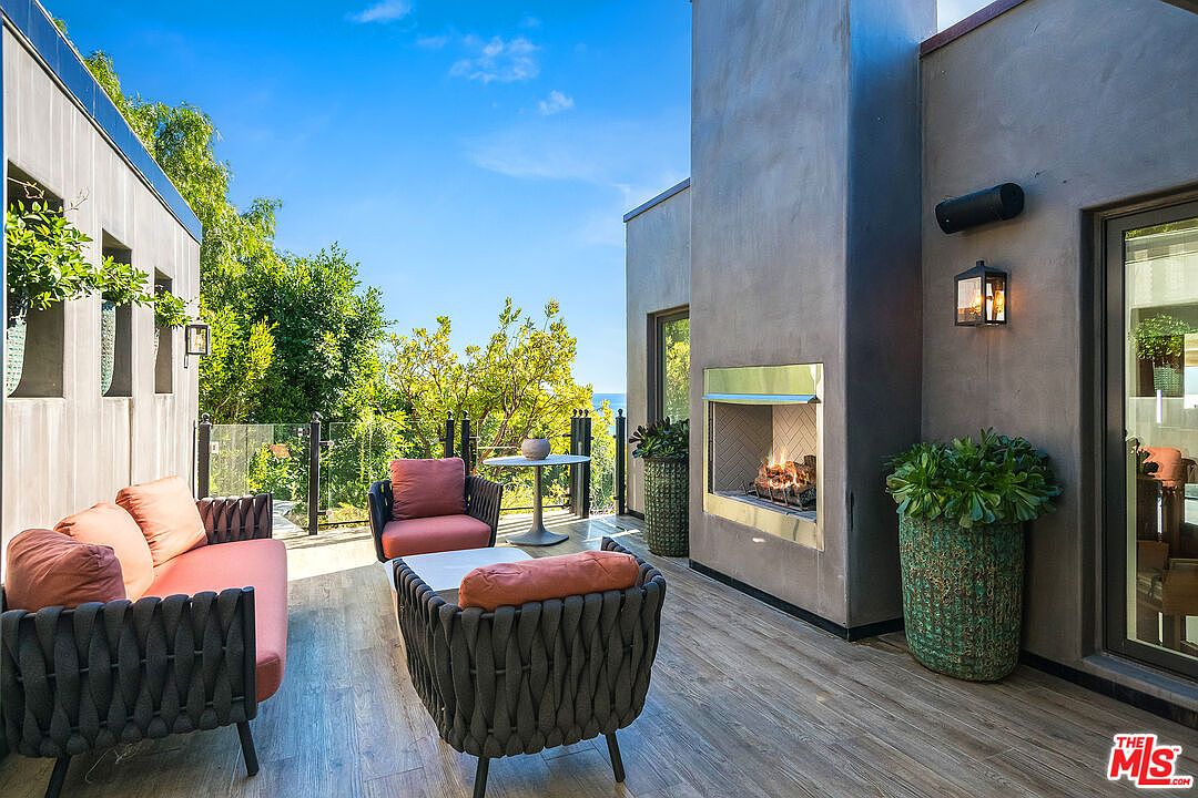 This modern outdoor patio features a sleek, built-in fireplace set into a dark stucco wall, creating a sophisticated focal point for relaxation. The space is furnished with contemporary woven lounge chairs and a sofa with plush orange cushions, all set upon warm-toned wood-look decking. A glass railing provides an unobstructed view of the lush greenery and distant ocean, enhancing the luxurious and serene atmosphere of this elevated terrace.