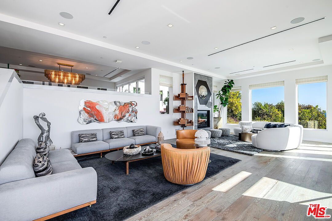 This expansive, open-concept living room features a sophisticated blend of modern design and warm textures, highlighted by a large grey sectional and two curved tan leather armchairs. The space is anchored by a dark area rug and a striking abstract art piece, while floor-to-ceiling windows provide abundant natural light and scenic views. A unique, tiered wooden shelving unit stands beside a contemporary fireplace, creating a focal point that exudes luxury and comfort.