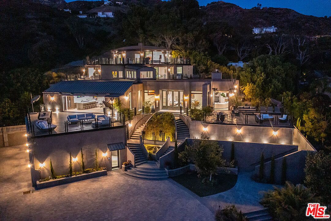 This stunning aerial night shot captures a multi-level luxury estate nestled against a hillside, showcasing its sophisticated architectural design and expansive outdoor living spaces. The property features a grand central staircase leading to the main entrance, multiple illuminated terraces, and a circular upper-level structure that offers panoramic views. The warm glow of exterior lighting highlights the modern textures and elegant landscaping, creating an inviting and opulent atmosphere.