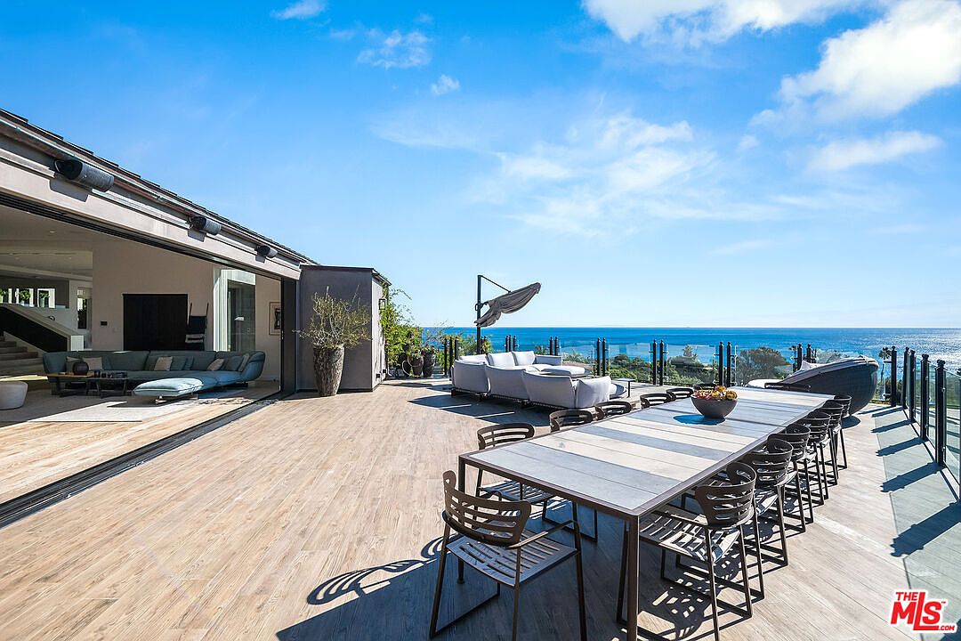 This expansive, modern outdoor deck offers a seamless transition from the indoor living area through large, retractable glass doors. The space features a long dining table set for outdoor entertaining and a comfortable lounge area, all overlooking a breathtaking, unobstructed view of the ocean. The clean lines, wood-look decking, and minimalist furniture create a sophisticated, high-end coastal living atmosphere.