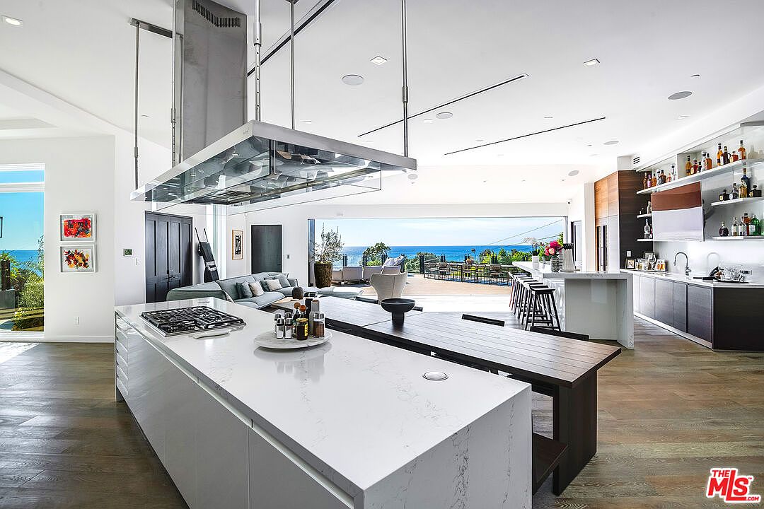 This expansive, modern open-concept kitchen features a massive white quartz waterfall island with an integrated gas cooktop and a sleek, suspended stainless steel range hood. The space seamlessly transitions into a dining area and living room, all framed by floor-to-ceiling glass walls that offer a breathtaking, unobstructed view of the ocean. The design utilizes a sophisticated palette of warm wood flooring, dark cabinetry, and minimalist lighting, creating a luxurious and airy atmosphere perfect for high-end coastal living.