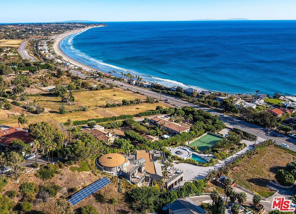 This stunning aerial view captures a sprawling coastal estate nestled between a scenic highway and the Pacific Ocean. The property features a unique architectural design with a circular roof section, a private tennis court, and a swimming pool, all surrounded by lush landscaping and open terrain. The expansive perspective highlights the prime location, offering direct proximity to the beach and panoramic ocean views.