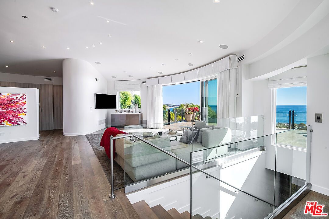 This bright and airy living room features a contemporary open-concept design with light wood flooring and a glass-railed staircase leading to a lower level. A plush, light-colored sectional sofa faces a large television and a sweeping wall of floor-to-ceiling windows that provide a stunning, unobstructed view of the ocean. The space is accented by a vibrant abstract painting and minimalist decor, creating a sophisticated and serene coastal atmosphere.