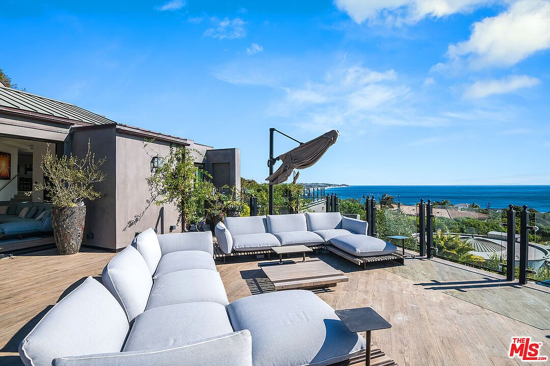 This expansive, modern outdoor deck offers a luxurious lounge area with plush, light-gray sectional sofas arranged for optimal ocean views. The space features a sleek, minimalist design with wood-look decking, a large cantilevered umbrella, and a glass railing that seamlessly connects the living area to the stunning coastal horizon. It is an ideal setting for high-end outdoor entertaining, capturing a serene and sophisticated atmosphere.