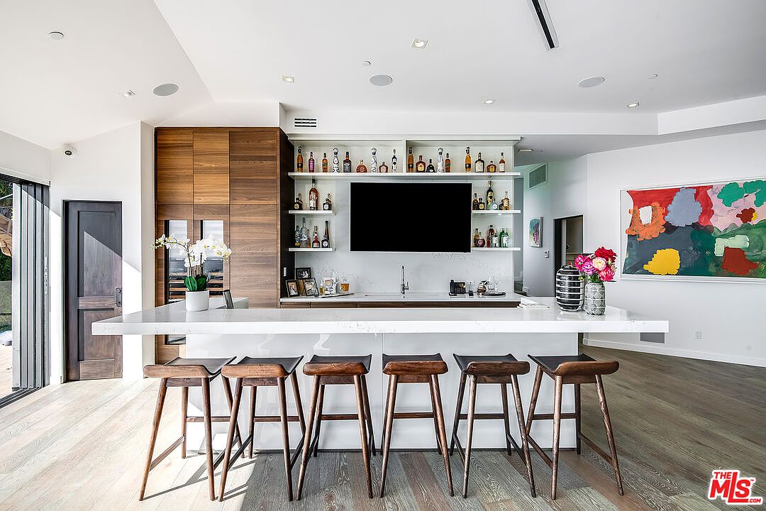 This modern, high-end kitchen features a sleek white quartz island with six dark wood bar stools, serving as a central focal point. Behind the island, a built-in wet bar area includes open shelving stocked with glassware and spirits, a mounted television, and a sink, all set against a clean, minimalist backdrop. To the left, warm wood cabinetry integrates a wine refrigerator, while a vibrant abstract painting adds a pop of color to the right, creating a sophisticated and inviting entertainment space.