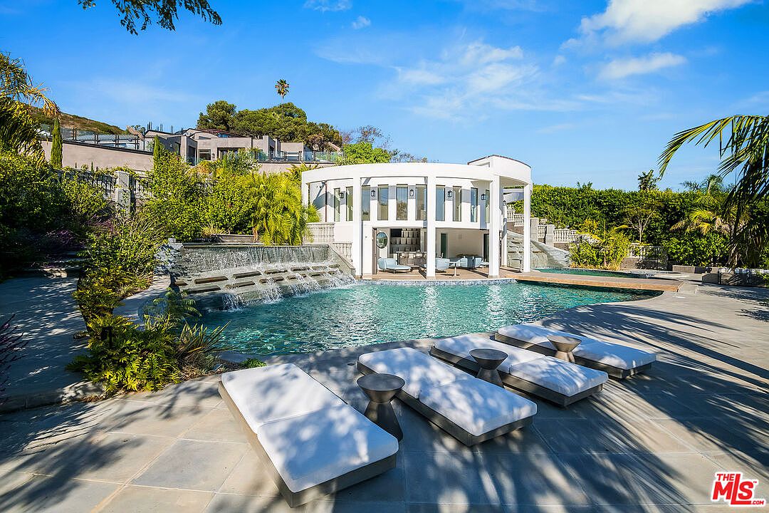 This luxurious outdoor space features a large, custom-shaped swimming pool with a tiered stone waterfall feature cascading into the water. A modern, circular white pavilion with floor-to-ceiling windows sits poolside, complemented by a stone patio furnished with sleek lounge chairs. The setting is lush and private, surrounded by mature landscaping and palm trees, creating a serene and high-end resort-like atmosphere.