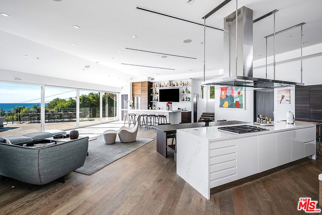 This expansive, open-concept living space seamlessly blends a modern kitchen with a bright, airy living area. The kitchen features a large white waterfall-edge island with a built-in cooktop, complemented by sleek cabinetry and a stainless steel range hood. Large floor-to-ceiling glass sliding doors open to a spacious deck, offering a stunning panoramic view of the ocean and creating a perfect indoor-outdoor living experience.