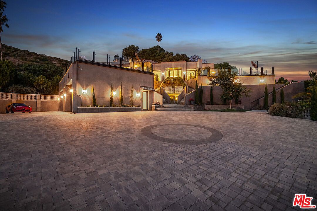 This stunning luxury estate features a grand, paved motor court leading to a multi-level architectural design with modern, clean lines. The exterior is illuminated by warm, ambient lighting that highlights the stone facade, tiered landscaping, and a prominent central staircase leading to the main entrance. A sleek red sports car is parked to the left, emphasizing the high-end, exclusive nature of the property at dusk.