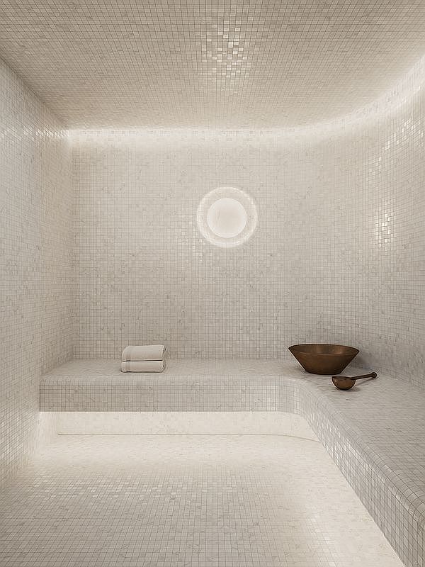 This image showcases a luxurious, minimalist steam room or hammam, entirely clad in small, light-toned mosaic tiles that create a seamless and serene aesthetic. A curved built-in bench provides seating, accented by soft under-bench lighting, while a circular wall light adds a warm, focused glow to the space. A bronze bowl and ladle rest on the bench, emphasizing the spa-like atmosphere and high-end design.