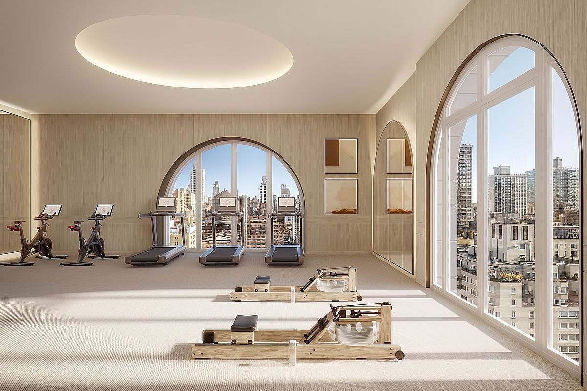 This sophisticated, high-end home gym features a minimalist aesthetic with light wood-toned walls and plush carpeting. The space is equipped with professional-grade exercise equipment, including stationary bikes, treadmills, and wooden rowing machines, all positioned to take advantage of the expansive, arched floor-to-ceiling windows that offer panoramic city views. The room is bathed in soft, natural light, creating an airy and serene atmosphere perfect for a luxury fitness experience.