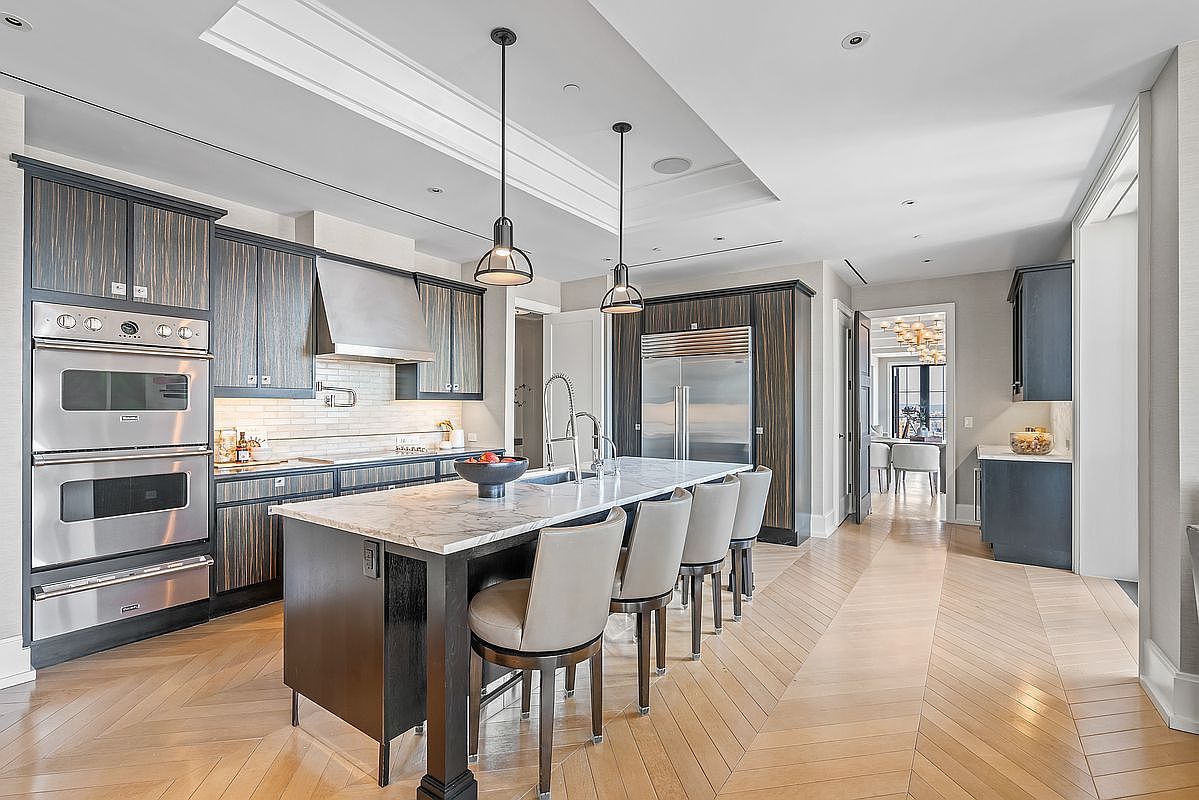 This sophisticated, high-end kitchen features dark, vertically-grained cabinetry paired with a large marble-topped island and light-toned chevron hardwood flooring. The space is anchored by professional-grade stainless steel appliances, including a double wall oven and a built-in refrigerator, all illuminated by elegant pendant lighting. The perspective is a wide-angle shot from the kitchen island looking toward the dining area, emphasizing the open-concept layout and luxurious finishes.