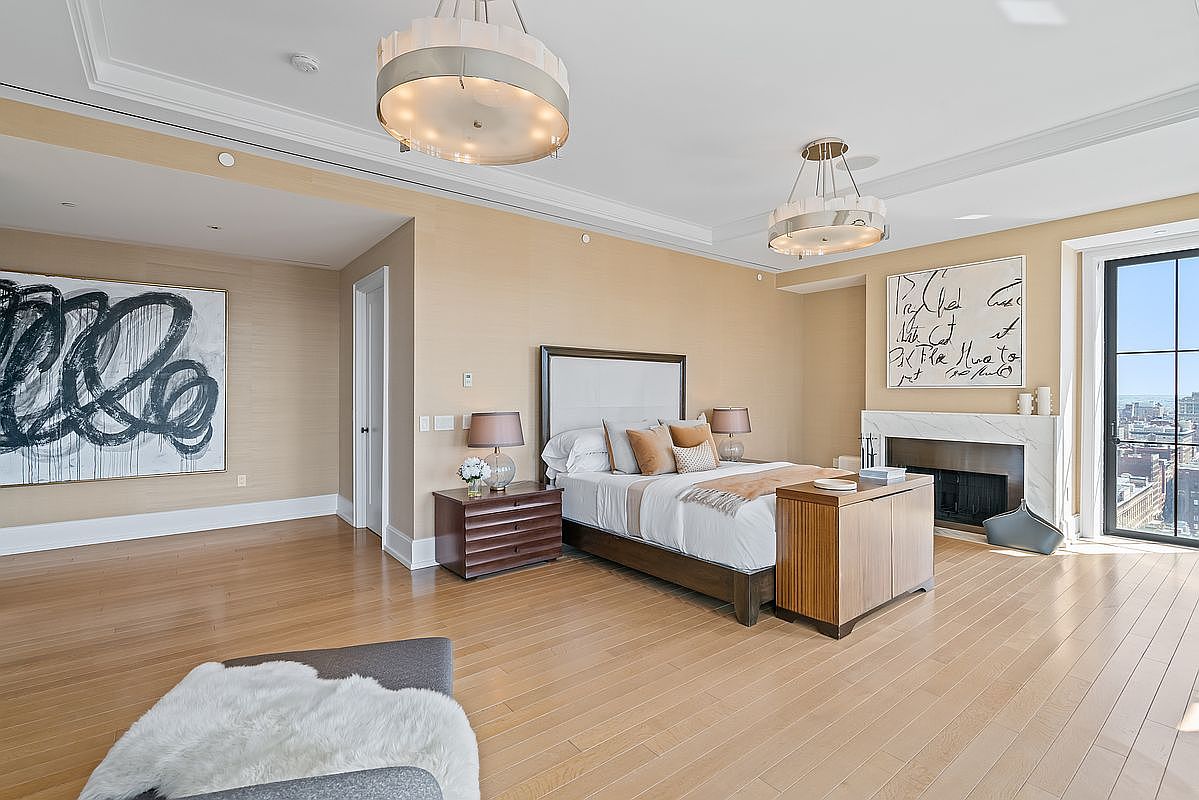 This spacious primary bedroom features a sophisticated, neutral color palette with light wood flooring and textured beige walls. The room is anchored by a large bed with a modern headboard, flanked by dark wood nightstands and elegant pendant lighting, while a fireplace with a marble surround and abstract artwork adds a touch of luxury. Large windows provide an expansive city view, creating an airy and high-end atmosphere.
