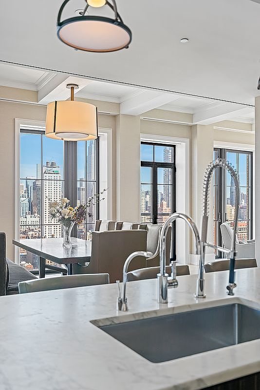 This perspective captures a sleek, modern kitchen island with a marble countertop and a professional-grade faucet, looking out toward a bright dining area. Large floor-to-ceiling windows frame a stunning urban skyline, while elegant pendant lighting adds a sophisticated touch to the open-concept space. The clean lines and neutral color palette create an airy, high-end atmosphere perfect for luxury city living.