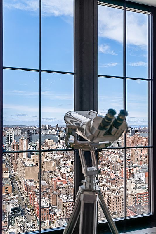 This image captures a sophisticated interior perspective looking out through a large, floor-to-ceiling window that frames a breathtaking panoramic view of a dense urban cityscape. In the foreground, a polished, professional-grade telescope on a tripod stands as a focal point, suggesting a luxury lifestyle and an appreciation for the surrounding skyline. The dark window frames contrast sharply with the bright, expansive blue sky and the intricate details of the city buildings below, creating a cinematic and aspirational atmosphere.