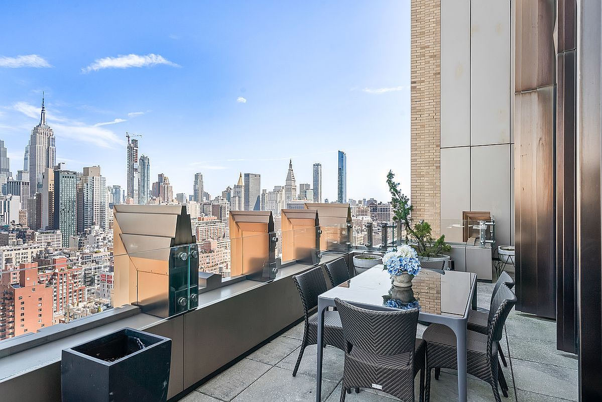This high-rise balcony offers a stunning, panoramic view of the New York City skyline, featuring the iconic Empire State Building. The space is furnished with a sleek, modern dining set, perfect for outdoor entertaining against an urban backdrop. The combination of glass railings, metallic accents, and the expansive cityscape creates a sophisticated and luxurious atmosphere.