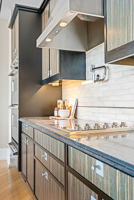 This modern kitchen features sleek, dark wood-grain cabinetry with a vertical striped pattern, paired with a light-colored subway tile backsplash and a polished stone countertop. A stainless steel range hood hangs above a built-in cooktop, while a pot filler faucet adds a functional, high-end touch to the workspace. The perspective is a close-up, eye-level shot that emphasizes the sophisticated textures and clean lines of the contemporary design.