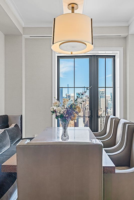 This elegant dining area features a sleek, modern table paired with upholstered chairs and a built-in bench, creating a sophisticated and intimate atmosphere. A large, contemporary drum pendant light hangs above the table, while floor-to-ceiling windows offer a stunning, expansive view of the city skyline. The neutral color palette and clean lines contribute to a refined, high-end urban aesthetic.