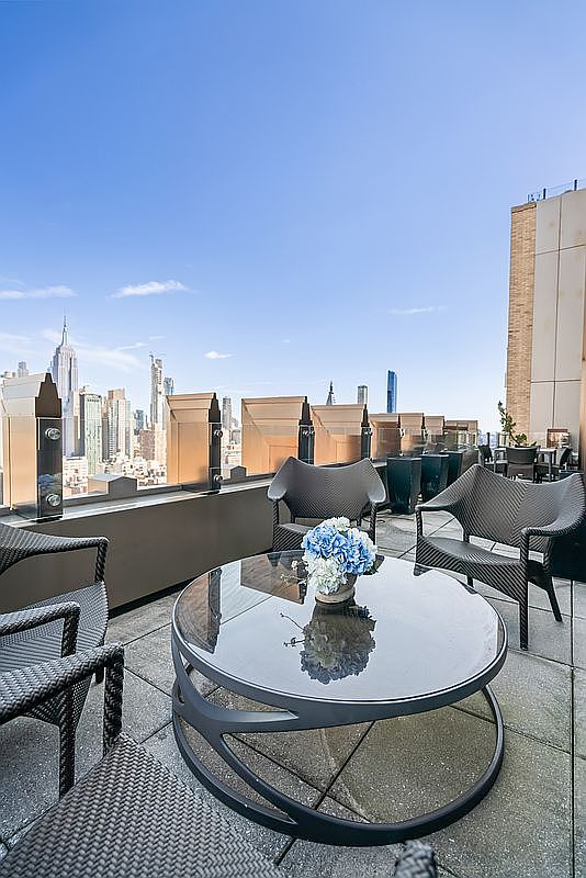 This high-rise rooftop terrace offers a sophisticated outdoor living space with a panoramic view of the New York City skyline, including the iconic Empire State Building. The area is furnished with modern, dark wicker-style seating arranged around a sleek, circular glass-topped coffee table adorned with a vibrant blue hydrangea centerpiece. The clean, minimalist aesthetic and elevated perspective create an atmosphere of luxury and urban tranquility.