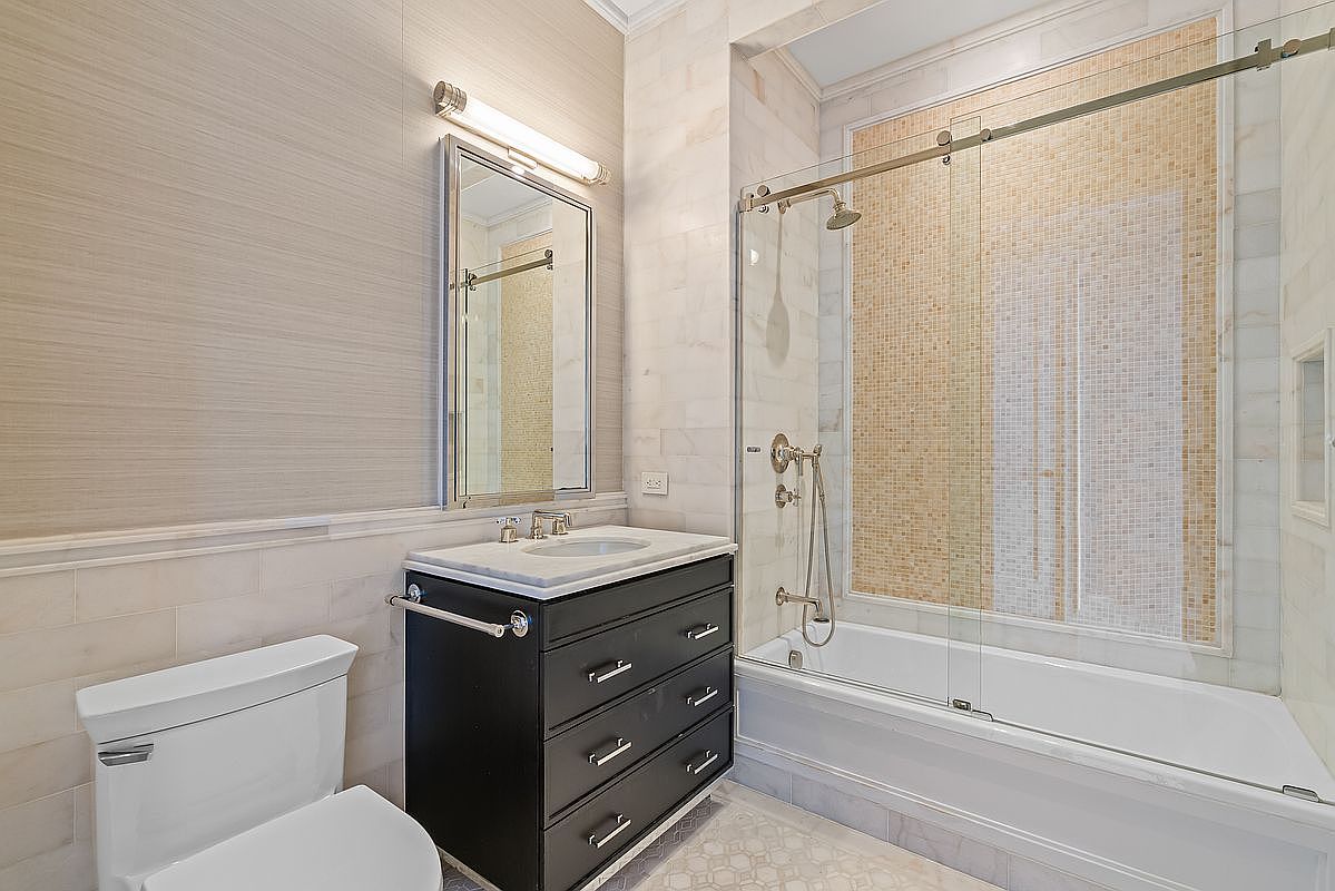 This elegant bathroom features a sleek black vanity with a marble countertop, paired with a modern glass-enclosed shower and tub combination. The walls are adorned with a mix of textured wallpaper and light-toned subway tiles, while a mosaic tile accent wall adds visual interest to the shower area. The space is well-lit by a contemporary vanity light, creating a clean and sophisticated atmosphere.