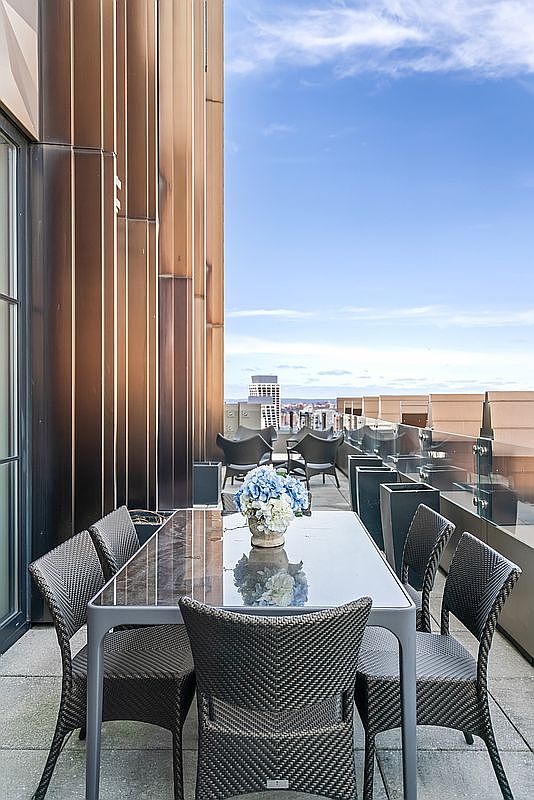 This high-rise balcony features a sleek, modern dining set with a glass-topped table and woven chairs, perfectly positioned for outdoor entertaining. The space is framed by striking, vertical copper-toned architectural panels and a glass railing that offers an expansive, unobstructed view of the city skyline. The overall atmosphere is sophisticated and urban, highlighting a premium outdoor living area.