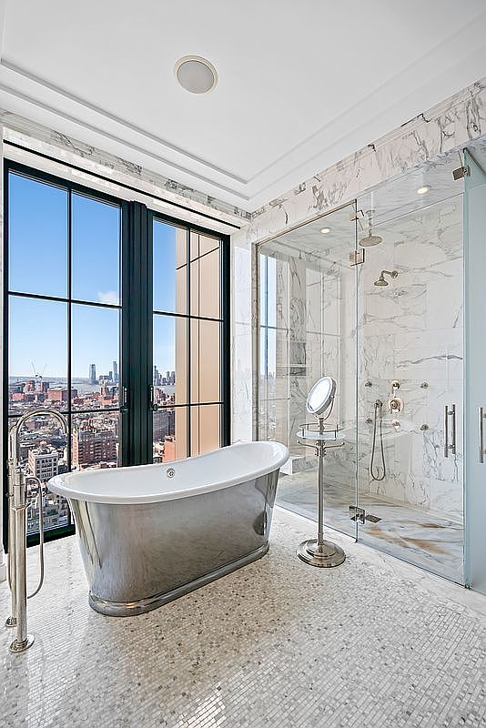 This luxurious primary bathroom features a striking metallic freestanding soaking tub positioned next to floor-to-ceiling windows that offer a breathtaking panoramic city view. Adjacent to the tub is a spacious, glass-enclosed walk-in shower clad in elegant white marble, complemented by a matching marble mosaic tile floor. The space exudes a sophisticated, high-end aesthetic, blending modern architectural design with opulent materials.