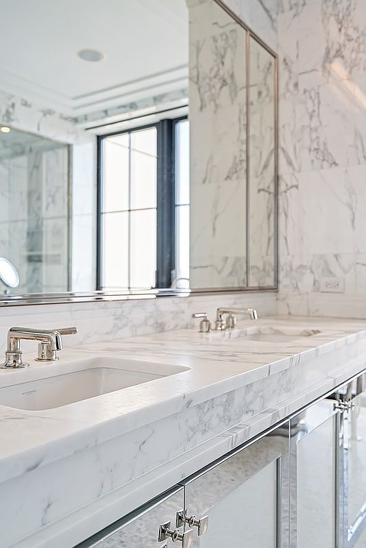 This luxurious primary bathroom features a double vanity with a polished marble countertop and integrated sinks, complemented by elegant chrome fixtures. The walls are clad in matching marble tile, creating a seamless and sophisticated aesthetic, while a large mirror reflects the bright, natural light from the nearby window. The overall impression is one of high-end refinement and clean, modern design.