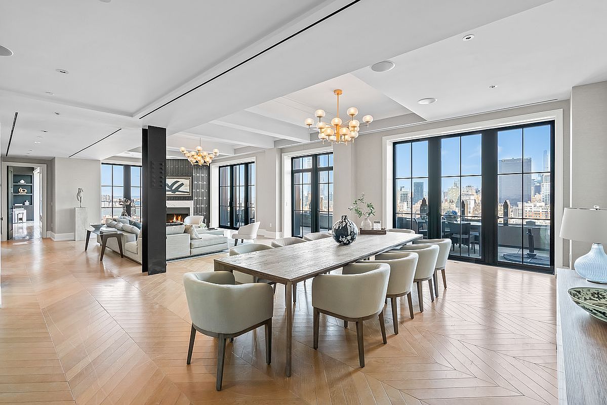 This sophisticated open-concept dining area features a long, rustic wooden table surrounded by elegant, light-toned upholstered chairs, set against a backdrop of expansive floor-to-ceiling windows that offer a stunning urban skyline view. The space is illuminated by a modern, multi-globe chandelier and flows seamlessly into a cozy living area with a fireplace, all tied together by rich, herringbone-patterned hardwood flooring. The overall aesthetic is one of refined luxury, emphasizing natural light and a spacious, airy atmosphere.